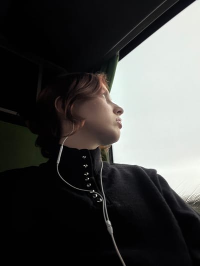 An individual with reddish-brown, wavy hair is observed in profile, seated inside a vehicle in Moineville, France, looking out a window to the right. The person wears a dark, high-collared garment with a vertical line of metallic stud-like fasteners on the left side of the collar. White wired earbuds are connected to the person's left ear, with the wire extending downward over the clothing. A silver hoop earring is visible on the left ear. The upper left portion of the frame shows the dark interior ceiling of the vehicle, with a green fabric element visible on the far left. The window on the right reveals an overcast, light grey sky, with visible rain streaks or condensation on the glass surface. The individual's gaze is directed upwards and outwards, towards the exterior view.