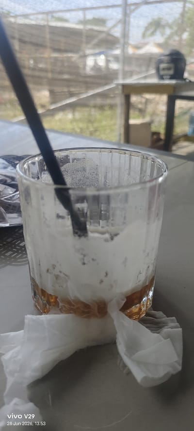 A close-up shot captures a nearly empty glass with a black straw and white foamy residue, sitting on a table. The glass contains a small amount of amber-colored liquid at the bottom. Two crumpled white paper napkins are placed in front of the glass. In the background, a blurred view of an outdoor area with some structures and vegetation is visible. The scene is set in Gampongpaya, Indonesia.