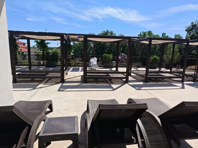 The image depicts an outdoor terrace or patio area in Starosel, Bulgaria, featuring a series of dark brown, rectangular wooden pergola structures with light-colored fabric canopies overhead. Underneath each pergola is a raised platform, likely intended for lounging. In the foreground are several dark brown wicker-style lounge chairs and a small matching side table. The terrace is paved with light-colored concrete. In the background, trees and greenery are visible, along with glimpses of buildings with red-tiled roofs. The sky is blue with scattered white clouds. A bird is seen in flight near the upper left corner of the image. One of the pergolas contains a covered chair. The scene suggests a resort or relaxation area.