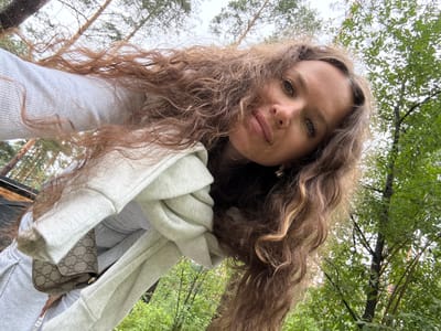 A low-angle, close-up selfie captures a young Caucasian woman with long, curly brown hair and green eyes looking towards the camera. She is wearing a light gray hooded sweatshirt, with a white scarf draped over her shoulder, and a small brown designer handbag with a monogram pattern is visible. The background consists of lush green trees and a cloudy sky, suggesting an outdoor setting likely in the city of Nizhnekamsk, Russia, possibly during the summer or early autumn. The perspective of the shot, taken from below, emphasizes her face and hair against the natural backdrop.