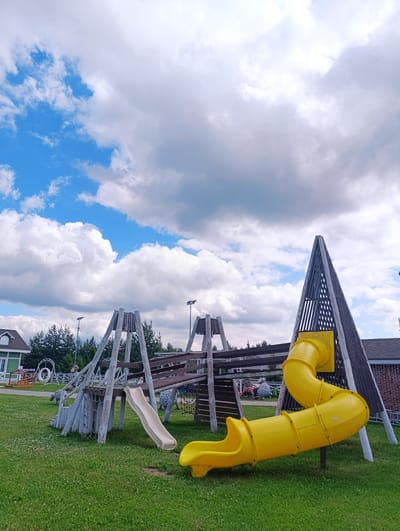 The image depicts an outdoor playground located in the city of Ivanovskoe, Russia. In the foreground, a vibrant yellow, curved slide descends from a wooden play structure onto the grassy ground. To the left, a smaller, cream-colored slide is attached to another section of the play area, which features wooden supports and a rope net. The play structure is made of weathered wood and appears to be quite large, with multiple levels and platforms. In the background, a distinctive triangular wooden structure with vertical slats stands prominently. To the far left, a green building with white trim and a visible roofline is partially visible. A dense line of green trees forms the backdrop behind the playground. The sky above is a bright blue, dotted with fluffy white clouds, suggesting fair weather. The ground is covered in green grass with small white flowers scattered throughout. There are no people visible in the image.
