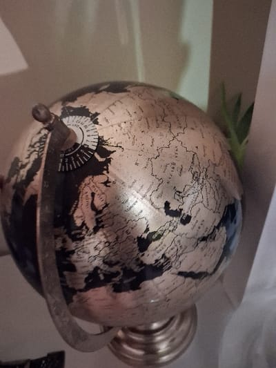 A globe with a golden landmass and dark seas sits in its stand. Detailed black lines outline continents and countries. A white circular dial with numbers is affixed to the globe, near the base. A metallic ring encircles the sphere. The stand appears to be made of a similar metal. The globe rests on a light-colored surface, with hints of greenery and white fabric in the background. The lighting emphasizes textures and creates highlights on the golden surface.