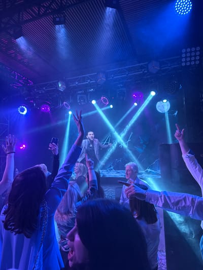 The image captures an indoor live performance event in Редант, Russia. A male performer with dark hair, wearing a grey jacket over a dark shirt, stands center-stage, mid-action with arms spread and mouth open, indicative of singing or engaging the audience. He is positioned in front of a large screen or projection displaying a blue-toned, possibly lunar, landscape with prominent white stylized Cyrillic letters "А А". Above the stage, a truss system supports numerous blue and purple LED lights and spotlights, casting visible beams through a hazy atmosphere. The audience occupies the foreground and midground, largely obscured but demonstrating active participation: multiple hands are raised, some individuals are pointing towards the stage, and one person holds a smartphone upward, likely recording. A grey-haired man in a light blue shirt is visible in the mid-right, looking towards the stage. A large dark speaker cabinet is situated on the right side of the stage. The venue's ceiling exhibits a corrugated metal structure. Confetti or small reflective debris is scattered on the floor in the immediate foreground.