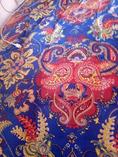 The image displays a close-up view of a blue textile with a paisley pattern. The pattern features swirls and stylized floral motifs in red, yellow, and green. A white USB cable is visible in the upper left corner, partially resting on the fabric. The lighting suggests an indoor environment. No specific location in Vostochnoe, Kyrgyzstan, can be identified from the provided image.