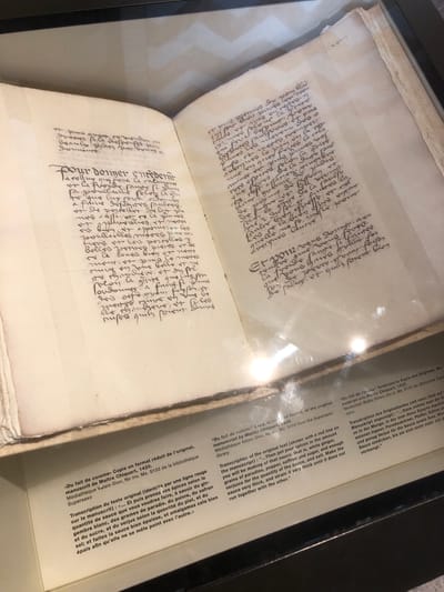 A photograph shows an open book displayed in a glass case in a museum or gallery setting. The book contains handwritten text in a dark ink, filling both pages with dense script that appears to be old French. The text is difficult to read in detail due to the handwriting style and possible degradation of the ink. To the right of the book, under the glass, is a typed transcript of the original text in French, German, and English.
No identifying information relates to Veytaux, Switzerland.