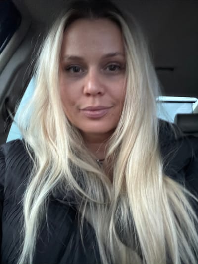 The image displays a close-up, front-facing portrait of a female individual with fair skin and long, straight, light blonde hair parted centrally. Her eyes are dark, and her facial expression is neutral. She appears to be wearing a dark, possibly black, garment with a pleated texture visible on her shoulders and chest. A thin, dark necklace is present around her neck. The background indicates the interior of a vehicle, with a dark headrest visible on the right side and a lighter, diffused area suggesting a window on the left. The scene is situated in Gol'yany, Russia.