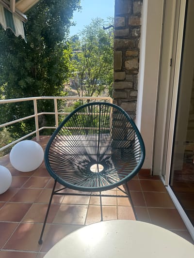 The image shows a balcony in Ospedaletti, Italy. A teal-colored woven chair is placed on the tiled floor, with two large white spherical objects to its left. A white table is partially visible in the foreground. Beyond the balcony railing, lush green trees and vegetation are visible, suggesting a natural, possibly hilly landscape. The sky is clear and blue. The architecture includes a stone pillar and a white wall next to a sliding glass door. The scene appears to be from a residential property, likely a home or hotel, given the balcony setting and outdoor furniture.