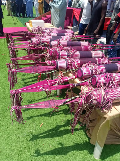 The image shows a table covered with multiple ceremonial drums and spears, likely in Dilpurtu, Nigeria. The drums are cylindrical with purple and white geometric patterns, adorned with matching tassels. The spears have purple, leaf-shaped blades and decorated shafts. A group of people stands in the background, some wearing formal attire. The scene takes place on a green, grassy surface, with a pink rug and a draped table also visible. Some people are observing and talking.