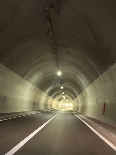 The image depicts the interior of a tunnel, likely a highway tunnel, as indicated by the asphalt road with white lane markings. The tunnel is illuminated by overhead lights, creating a sense of depth as they recede into the distance. The walls and ceiling of the tunnel appear to be constructed from concrete, with some textured or irregular surfaces visible. There is a red sign or banner visible on the far wall with text in what appears to be Cyrillic or Latin script. The perspective suggests the photo was taken from a moving vehicle. The location is identified as Bukovica, Montenegro, which is known for having several significant tunnels, including the Klisur and Vjetrenik tunnels which are part of the main road connecting Podgorica to the coast.