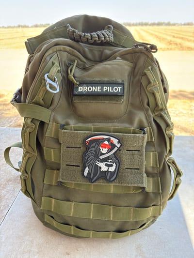 An olive drab tactical backpack stands upright on a light-colored tiled surface in an outdoor setting within the Merhavim Regional Council, Israel. The backpack features extensive MOLLE webbing across its front and sides. A rectangular black patch with white text reading "DRONE PILOT" is affixed to a velcro panel on the upper front. Directly below, on a separate MOLLE-compatible panel, is a rubber patch depicting a grim reaper figure. This grim reaper wears a black hooded cloak, has a white skeletal head with red glowing eyes and a red blindfold, and holds a white-bladed scythe in its left hand while operating a black drone remote controller with its skeletal right hands. A silver carabiner clip is attached to a strap on the upper left side of the backpack. A tightly woven olive drab paracord handle is integrated into the top carrying strap. Bright direct sunlight illuminates the scene from the right, casting distinct shadows on the tiled surface to the left of the backpack. The background reveals an arid agricultural landscape composed of fields with dry, cut crops extending towards a hazy horizon under a clear sky.