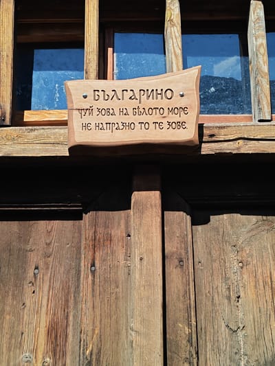 This image captures a close-up of a traditional wooden facade, likely belonging to a house or building in the village of Kovachevitsa, Bulgaria. The primary subject is a carved wooden sign affixed to the structure, centered horizontally in the upper-middle portion of the frame.
The sign is made of light-colored, natural wood, approximately rectangular with undulating, organic edges, suggesting a handcrafted origin. It is secured to a darker wooden beam or frame by at least one visible metal stud on its left side. Engraved into the sign in Cyrillic script are three lines of text:
1. БЪЛГАРИНО
2. ЧУЙ ЗОВА НА БЯЛОТО МОРЕ
3. НЕ НАПРАЗНО ТО ТЕ ЗОВЕ.
This translates to:
1. BULGARIAN (as an address)
2. HEAR THE CALL OF THE WHITE SEA
3. IT DOES NOT CALL YOU IN VAIN.
(The "White Sea" refers to the Aegean Sea in historical Bulgarian context.)
Above the sign, a section of two distinct, rectangular window panes is visible. The blue-tinted glass of these windows reflects a bright blue sky with fragmented white clouds or distant landscape features. These window panes are framed by robust, dark brown wooden structural elements, consistent with traditional Bulgarian Revival period architecture found in Kovachevitsa.
Below the sign, a section of a dark brown wooden wall or door is depicted, constructed from vertically oriented planks. These planks exhibit pronounced wood grain, knots, and variations in tone, indicating weathering and age. Several small, circular metal nails or fasteners are visible on the surface of these planks. A deep, sharp shadow is cast by the upper architectural elements and the sign itself onto the lower wooden surface, suggesting strong, direct sunlight from an elevated position. No people are present in the scene.