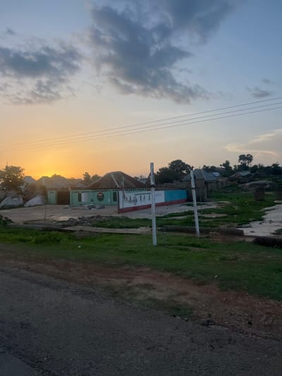 This detailed image captures a rural or semi-urban scene in Ribina East, Nigeria, at either sunrise or sunset, indicated by the vibrant orange glow on the horizon. The sky transitions from a bright orange at the bottom to a pale blue at the top, interspersed with scattered dark grey clouds. Three horizontal power lines traverse the upper portion of the sky.

In the mid-ground, a prominent single-story building with a light green facade and a dark, ridged roof is visible. Painted on its wall is "TORO LOCAL GOVERNMENT PARTY SECRETARIAT," flanked by circular logos for "ADC" and "APC." To the right of this building, another structure features a wall painted in light blue and red, displaying text that reads "DAMANAHAN WUNTI FOR REPS APC" above images of individuals. Large, irregular rock formations and boulders are scattered around the buildings, particularly on the left side of the scene.

The immediate foreground consists of a dark, textured ground, possibly a road or wet dirt, leading into an area of green grass and patches of reddish-brown earth. Two tall, slender white poles stand vertically in the center-right mid-ground. No people are visible in the frame. Trees and additional buildings with dark roofs are sparsely distributed in the background, set against the rocky landscape.