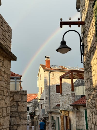 A rainbow arches across a pale gray sky above a narrow, stone-lined street in Primošten, Croatia. The street is flanked by multi-story buildings constructed of light-colored stone, with red-tiled roofs. A black outdoor lantern with a conical shade hangs from a rusty bracket mounted on the wall of a building on the right. The bracket itself is a horizontal wooden beam with three ceramic insulators, each topped with a small, dark brown cap. Solar panels are visible on the roof of a building in the middle ground, and a wooden pergola with a light-colored canopy extends from another building. Two people, one in a blue shirt, walk away from the camera down the street, while two others in white shirts are further ahead. The stone walls of the buildings create a textured, rustic aesthetic.