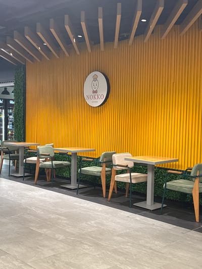 The image depicts an interior view of a cafe or restaurant, possibly in Amstetten, Austria, judging by the architecture and the presence of a "NOKKO Kitchen" sign. The walls are lined with horizontal yellow paneling. Parallel wooden slats are arranged on the left side of the frame, with embedded lighting fixtures. A round sign with the "NOKKO Kitchen" logo is visible on the wall.
Several tables and chairs are aligned along the right side of the frame. The chairs have light green cushions and wooden frames. Decorative green foliage is present behind the chairs. The floor is paved with light-colored tiles. A glimpse of a shopping area with various storefronts can be seen through a distant window.