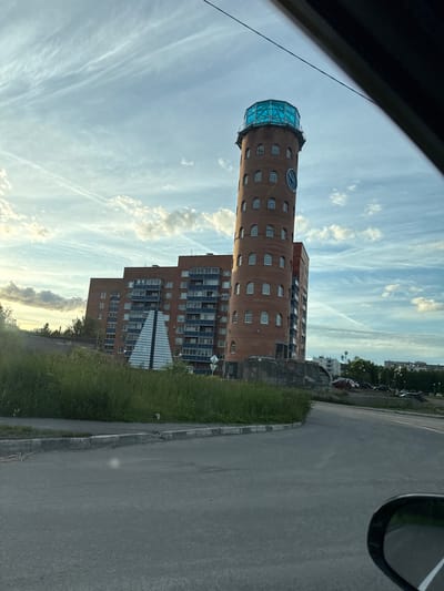 A brick building with a cylindrical tower and a glass dome on top stands next to a multi-story apartment building. The tower has a large clock face. The sky is blue with wispy white clouds, suggesting it is daytime. A paved road runs alongside the buildings, with a strip of grass and a curb separating it from the structures. In the background, more buildings and trees are visible. The image appears to be taken from inside a vehicle, with a portion of the car's interior visible in the upper left corner, possibly indicating the viewer is a passenger or driver. The overall scene depicts an urban or suburban environment in Kondopoga, Russia. The lighting suggests it could be late afternoon or early evening. There is no visible text in the image.