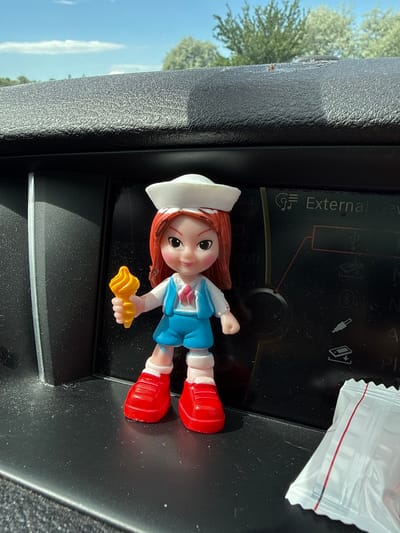 A brightly lit, slightly tilted photograph captures a small figurine placed on a textured, dark surface within a vehicle's interior. The figurine depicts a girl with reddish-brown hair, a white cap, a light blue top with white sleeves, and red boots. She has a gentle smile and is holding a yellow, shell-shaped object in her left hand.
The figurine rests on what appears to be a car's dashboard or center console. Behind it, a dark panel or screen displays various icons, including symbols for USB and Bluetooth, and the partially visible word "External" in white text. To the right of the figurine, a corner of a small white plastic packet with red and black striped edges is visible.
Through the car window on the left, a clear blue sky dominates the outdoor background, featuring a thin, elongated white cloud. Green foliage from trees is visible, suggesting an outdoor setting. The strong, direct light source indicates it is daytime with sunny weather. The scene is static, showcasing the figurine in this particular setting, located in Radinovo, Bulgaria.