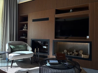 The image depicts a well-decorated living room. A light-colored armchair with two green throw pillows sits on a textured rug. Adjacent to the armchair is a black metal side table. In the background, a modern entertainment center features a large flat-screen television mounted above two recessed fireplaces. The left fireplace contains a set of fireplace tools, while the right fireplace is filled with a stack of logs and a small woven basket. Books and a remote control are placed on a circular black metal coffee table in front of the fireplaces. A tall, grey curtain hangs to the left of the armchair. The walls are paneled with dark wood, and the shelves above the television hold decorative items, including a small Buddha statue and a small sculpture. The overall ambiance is cozy and sophisticated, suggesting a living space in Bakovići, Montenegro.