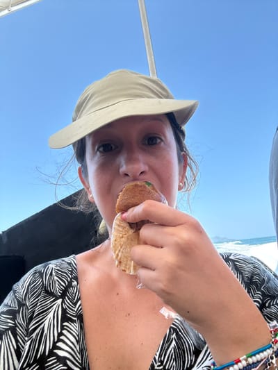 An outdoor, eye-level image captures a female individual, approximately middle-aged, aboard a vessel under a clear blue sky, identified as being in the city of Pak Nam, Thailand. The individual is centered in the frame, wearing a light-colored sun hat and a black and white patterned V-neck top with a leaf motif. She is actively consuming a round, brown-crusted food item, holding it in her right hand with a partial bite taken. The food is cradled in a fluted light-brown paper wrapper, partially peeled down. Green contents are visible within the bite mark. The individual wears small, circular stud earrings and multiple colorful beaded and metal bracelets on her left wrist. In the background, the sky is uniformly blue and clear. To the right, open water with visible white-capped waves indicates a body of water such as an ocean or sea, with a faint, distant landmass on the horizon. Elements of the vessel's structure, including a dark panel on the left and a white support pole on the upper right, are visible.