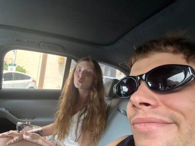 Two individuals are in a car, likely taking a selfie. One person wears sunglasses and is positioned closer to the camera. The other person is in the background. The scene is likely in Hongkou District, China, given the writing on the sign visible through the car window, which is likely Chinese script. A light-colored building is visible outside, and another vehicle is parked nearby.