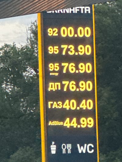 The image shows a digital price board at a gas station, likely in Shatava, Ukraine, as indicated by the presence of Cyrillic text. The board displays prices for various fuel types and other services. From top to bottom, the displayed information is:
- At the very top, partially visible white text reads "KINHFTA" followed by what appears to be the company name.
- The first fuel price listed is "92 00.00".
- Below that is "95 73.90".
- Underneath, "95 energy 76.90".
- Then, "ДП 76.90". "ДП" likely stands for Diesel.
- Next is "ГАЗ 40.40". "ГАЗ" means Gas.
- The final fuel price is "AdBlue 44.99".
- At the bottom, there are icons for a coffee cup, cutlery (fork and knife), and "WC".
The background consists of dark green trees and a cloudy sky. The price board itself is black with yellow illuminated digits and white text, bordered by a bright yellow vertical stripe. The overall scene is captured at what appears to be daytime, with natural light illuminating the surroundings.