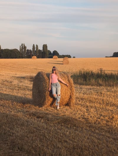 In Alvimare, France, a woman with blonde hair and sunglasses stands in a field of harvested wheat during sunset. She is leaning against a large bale of hay, wearing a pink tank top and ripped blue jeans. The field stretches out behind her, dotted with several more hay bales and a line of trees in the distance. The sky is a soft blue with wispy clouds, and the setting sun casts a warm, golden light over the landscape.