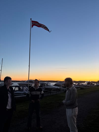 The image depicts an outdoor scene at dusk, likely in Tønsberg, Norway, with a vibrant sunset painting the sky in hues of orange and blue. A Norwegian flag flies prominently on a tall flagpole against the twilight sky. In the foreground, three individuals are visible, standing near a harbor filled with docked boats. The person on the far left is partially obscured and out of focus, wearing dark clothing and appearing to be talking. To their right, a man in a striped long-sleeve shirt and dark pants stands facing forward, his hands clasped in front of him. On the far right, another man, also in a striped long-sleeve shirt and light-colored pants, is seen in profile, looking towards the left. The background features a row of boats, suggesting a marina or harbor setting, with the water and distant land visible beyond. The overall atmosphere is calm and relaxed, possibly indicating a social gathering at the end of the day.