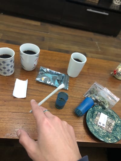 The image presents an indoor scene on a wooden table, likely in Beccar, Argentina. The composition shows various items arranged casually, alongside a person's hand.
In the foreground, a person's right hand, adorned with a ring featuring blue and gold bands, is visible. The hand is positioned near a white, hand-rolled cigarette, which appears to be a joint or blunt.
On the wooden table, there are three ceramic cups, each containing a dark liquid, likely coffee or tea. One cup is plain white, another is white with subtle red markings, and the third displays a grey and white diamond pattern.
Several objects related to cannabis use are scattered across the table. These include a clear plastic baggie filled with green leafy material (consistent with cannabis), a blue cylindrical grinder, and another dark blue cylindrical object that may also be a grinder or container. A small, crinkled metallic pouch and a folded piece of white paper are also present. A distinctive green ceramic plate, decorated with an embossed cannabis leaf pattern, holds a small white label. The text on this label is partially obscured and indistinct, but includes characters resembling "王" (Wang or king in Chinese) repeated, and possibly "JC" or "SC". In the upper left corner, a small decorative figurine is partially visible.
The scene depicts a casual setting, suggesting an activity involving both beverage consumption and the preparation or use of cannabis.