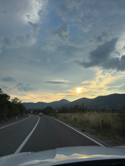The image depicts a scenic view from a moving vehicle on a winding road in Barutana, Montenegro. The time of day appears to be late afternoon or early evening, as evidenced by the low sun casting a warm glow through a partly cloudy sky. The sun's rays create a dramatic effect as they pierce through the clouds, illuminating the landscape and the sky with a soft, golden light.

The road is paved with asphalt and marked with a white line, suggesting it's a well-maintained route. To the right of the road, dry grass and vegetation line the embankment, typical of a Mediterranean or mountainous region. In the distance, rolling hills and mountains form the backdrop, their silhouettes softened by the atmospheric conditions.

Several vehicles are visible on the road, indicating it's a public thoroughfare. One car is ahead, and another is further down the road. A triangular traffic sign with a red border and a white cross symbol is posted on the right side of the road, warning of an intersection or hazard. The left side of the image is dominated by a vast expanse of clouds, ranging from dark, heavy masses to lighter, wispy formations, creating a sense of depth and dynamic weather.

The overall mood of the image is serene and picturesque, capturing the beauty of the Montenegrin landscape during a moment of transition in the day. The perspective from within a vehicle suggests a journey or a scenic drive, inviting the viewer to experience the visual richness of the location. There are no people clearly visible in the image.