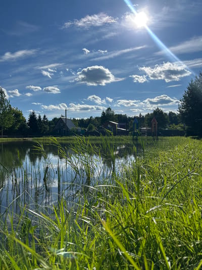In the city of Слава Снт, Russia, a body of water is framed by tall, green grass in the foreground, with reflections of the sky and surrounding trees visible on its surface. Across the water, a dense line of trees serves as a backdrop for several structures, including a house with a dark roof and a visible chimney, and a colorful playground with red and blue rocket-shaped structures. The sky above is a vibrant blue, dotted with fluffy white clouds and illuminated by the bright sun, which casts a strong lens flare. The overall scene depicts a peaceful, rural setting during daylight.