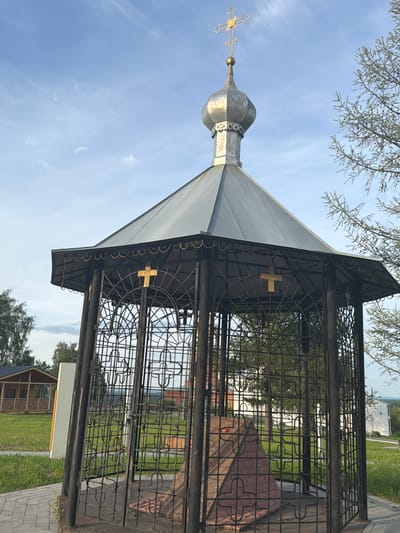 The image depicts a small, octagonal gazebo with a domed roof and a golden cross at its apex, located in Cherdyn, Russia. The gazebo is constructed of dark metal latticework, enclosing a stone monument or altar. The monument is pyramid-shaped and made of reddish-brown material, possibly brick or stone. There are golden crosses visible on the exterior of the gazebo's railings. The surrounding environment includes a paved pathway leading up to the gazebo, a well-maintained grassy area, and trees. In the background, a small wooden building with a porch is visible. The sky is clear with a few wispy clouds. The scene appears to be a religious site or a memorial.