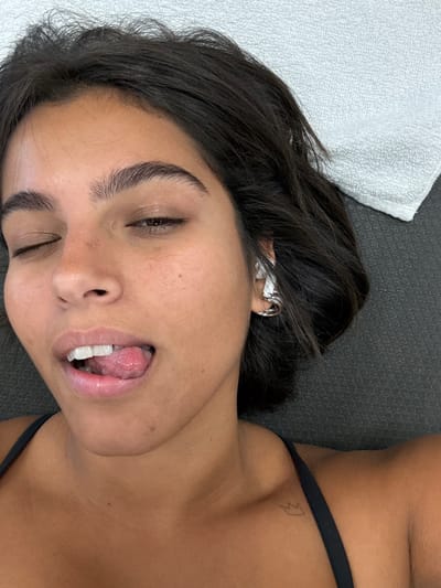 A female subject is depicted in a close-up portrait, lying on a dark, textured surface, likely a mat, with a white terry cloth towel positioned to her upper right. Her head is oriented towards the top of the frame. She possesses dark hair, dark eyebrows, and a medium skin complexion with visible freckles on her nose and left cheek. Her left eye is closed, and her right eye is partially closed. Her mouth is open, with her tongue extended and resting on her lower lip. A silver-toned ear cuff is present on her right ear. She is wearing a black top or sports bra, with one strap visible on her right shoulder. A small crown-shaped tattoo is visible on her right clavicle. The scene is located in Plainsboro Township, United States.