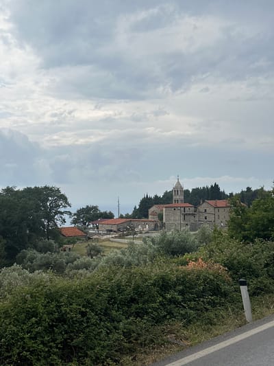 The image captures a serene, overcast daytime view of a village, likely Gornji Katun in Montenegro, nestled within a verdant, hilly landscape. The scene is dominated by traditional stone buildings, many featuring distinctive red-tiled roofs, set amongst dense green foliage.
A prominent stone church, characterized by its bell tower topped with a cross, stands out among the structures. The buildings appear to be old and well-integrated into the natural surroundings. No people or overt activities are visible, contributing to the tranquil atmosphere.
The foreground and sides of the image are filled with lush trees and thick bushes, including what appear to be olive trees. On the right, a portion of an asphalt road is visible, featuring a white lane marking. Alongside the road, a grey roadside marker stands, bearing some indistinct text that is not clearly legible.
The sky is largely covered by scattered, light grey clouds, indicating an overcast day with soft, even lighting, suggesting either late morning or early afternoon. The overall impression is one of peaceful rural life, showcasing the region's traditional architecture and natural beauty.