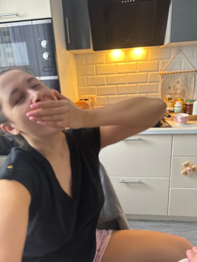 This image depicts an indoor scene within a kitchen in the city of Nikolina Gora, Russia. A single individual, an adult female, is positioned in the foreground. Her face is out of focus, with her eyes appearing closed or squinted, and her right hand, featuring pink painted nails, covers her mouth and nose. She is wearing a black v-neck short-sleeved top and light-colored, patterned shorts.

The background displays a modern kitchen environment. To the left, a built-in oven and microwave combination unit with a metallic finish and two circular control knobs is visible beneath a dark upper cabinet. In the center-right, a white subway tile backsplash is illuminated by two warm-toned under-cabinet lights. Above the backsplash, a dark range hood with a control panel and five small white indicator lights is mounted between dark grey upper cabinets. A light-colored countertop spans the scene. On the right side of the countertop, an orange, compact toaster-like appliance is present, alongside various jars, containers, and a pink cup. A light-colored fabric banner displaying the text "bee yourself" and a bee graphic hangs on the wall above the counter. The lower portion of the kitchen features white flat-panel cabinets and drawers with horizontal bar pull handles. A small decorative item resembling a floral or propeller shape with a yellow center is affixed to one of the drawer fronts. A light grey floor covering is partially visible at the bottom right.