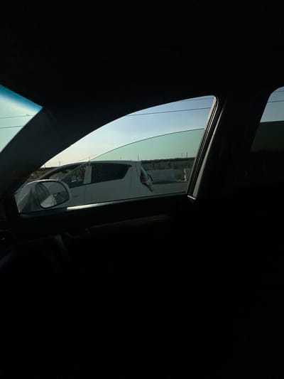The image shows a view from inside a vehicle looking out through the side window. The window is partially open, revealing a reflective surface that shows a distorted reflection of the surroundings. The interior of the car is dark, with the black trim of the window frame creating a strong contrast with the brighter exterior. Outside, a light-colored sky suggests it is daytime, possibly morning or late afternoon due to the low angle of the sun. The reflection in the window shows what appears to be a road or tarmac surface and some structures, possibly buildings or industrial elements. There is also a reflection of a side-view mirror mounted on the exterior of the vehicle. The overall impression is of a journey, with the focus on the interplay of light, reflection, and the surrounding environment as seen from within the car. The scene appears to be outdoors, with clear skies. The precise location cues from Istiqlol, Uzbekistan are not explicitly visible but the context suggests an urban or semi-urban environment.