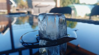 The image shows an ice cube melting on a dark, reflective surface, likely a table. The ice cube is translucent with visible air bubbles and frost patterns. It's partially submerged in a puddle of water that has formed around its base. The surface reflects the surrounding environment, showing blurred shapes of trees and buildings under a bright sky, suggesting it's daytime. In the background, out-of-focus elements include dark chair backs and what appears to be an outdoor setting. The lighting suggests a sunny day, possibly late afternoon due to the warm tones in the reflections. There is no visible text in the image. The scene evokes a sense of transient beauty and the passage of time as the ice melts. The close-up perspective focuses on the ice cube and its interaction with the melting process.