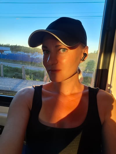 This is a close-up, vertically oriented, outdoor photograph taken of a young woman from the chest up. She is wearing a black tank top and a black baseball cap with a white brim. She also has black wireless earbuds in. The woman is looking towards the camera with light blue eyes and fair skin. She has blonde hair, which is visible sticking out from under her cap. The right side of her face and neck are illuminated by warm, golden sunlight, suggesting it is either sunrise or sunset. The lighting casts a strong shadow across the left side of her chest and shoulder.

The background shows a large window with a view of an outdoor scene. Through the window, a wooden deck or boardwalk is visible, along with greenery and what appears to be a body of water or a field. The sky is a clear, bright blue, with a few thin, horizontal lines that could be power lines. The overall impression is that the woman is inside a vehicle, possibly a train or a bus, with the sun shining in through the window. The mood is relaxed and possibly reflective, given the soft lighting and the woman's calm expression. No visible text is present in the image. The location context provided is Volkhov, Russia.