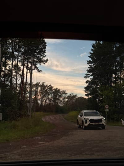 The image shows a white Haval SUV driving on a dirt road in Novoe Mart'yanovo, Russia. The road is wet and appears to have puddles. Tall trees line the left side of the road, and a power pole with a triangular sign is visible. On the right side of the road, there are more trees and a speed limit sign indicating 30 kilometers per hour. The sky is partly cloudy with hues of blue and pink, suggesting it is either sunrise or sunset. The image is taken from a low angle, possibly from inside a vehicle.