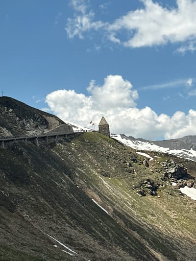 This is a landscape photograph taken from a low angle, looking up at a mountainous terrain under a bright blue sky with scattered white clouds. In the mid-ground, a stone tower with a pointed roof stands on a grassy slope dotted with patches of snow. Two flags are visible near the tower, one red and white, and another yellow and red. A paved road with a stone retaining wall and a wooden railing follows the contour of the mountain slope to the left of the tower. The mountainside is steep and covered with a mixture of green vegetation, rocks, and patches of snow. The foreground is dominated by the rocky and vegetated slope of the mountain. The scene appears to be in a high-altitude mountainous region.