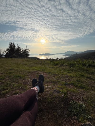 A first-person perspective photograph captures the lower legs of an individual resting at an elevated viewpoint in Vojnići, Montenegro. The subject's legs are extended and crossed at the ankles, clad in maroon ribbed leggings, white crew socks with two maroon horizontal stripes, and black hiking boots. The immediate foreground consists of sparse green grass, brown soil, and scattered small rocks. The midground features a grassy hillside, with a cluster of dark green pine trees visible on the left. The background presents a wide coastal panorama: a sun low on the horizon, positioned left of center, casts a bright, shimmering reflection across a body of water, likely a bay or the sea. Distant islands or landmasses are visible within the water, and undulating hills or mountains recede into the hazy distance on the right. The sky is largely covered by a textured pattern of numerous small, uniform cirrocumulus clouds against a pale blue expanse. The lighting suggests either sunrise or sunset.