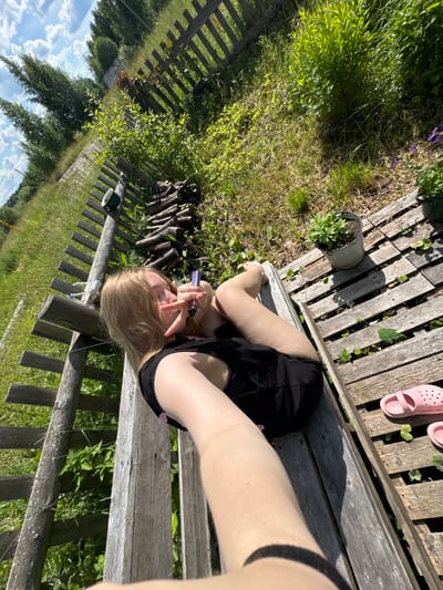 The image depicts a young person lying on a wooden platform in an outdoor setting. The platform appears to be constructed from weathered wooden planks. The person, with blonde hair and wearing a black tank top and shorts, is positioned on their back, facing the camera. They are holding a purple electronic cigarette in their right hand and making a "peace sign" with their fingers.
The environment surrounding the platform is characterized by lush green grass and vegetation, suggesting a rural or semi-rural location. A rustic wooden fence runs along the left side of the frame, made of vertical posts and horizontal slats. Beyond the fence, tall green trees are visible against a blue sky with scattered white clouds. To the right of the platform, there are several potted plants, one in a galvanized metal pot with green foliage. A stack of firewood is piled near the fence. Two pink Crocs-style sandals are placed on the wooden planks near the bottom right corner of the image. The overall scene suggests a sunny day, possibly in a backyard or a similar outdoor leisure space. The presence of a fence and the natural surroundings could indicate a location in the village of Ryabinino, Russia, known for its rural landscapes and wooden architecture.