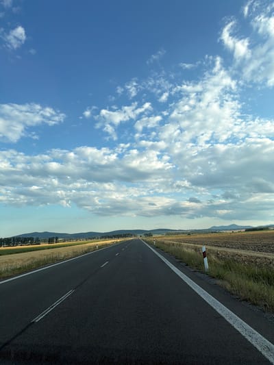 This is a photograph of a road in Kožuchov, Slovakia, with a clear sky filled with white clouds. The road is asphalt and has a white line marking the edge and a double white line in the middle, indicating a two-way street. The road appears to be in good condition.
To the right of the road, there is a field of dry grass and a patch of cultivated land, suggesting it is autumn or a dry season. The cultivated field is a light brown color. There are also a few white road markers with red reflectors on the right side of the road.
To the left of the road, there is a field of golden dry grass and then a line of trees. Further in the distance, there are rolling hills covered in a green forest, and beyond that, mountains are visible under the horizon.
The sky is a vibrant blue with scattered white clouds. Some clouds are large and puffy, while others are thinner and wispy. The lighting suggests it is daytime, possibly late afternoon, as the sun is not directly overhead and the light is somewhat softened.
There are no people or vehicles visible in the image, making it a tranquil landscape scene. The overall impression is one of open space and a journey through the Slovak countryside.