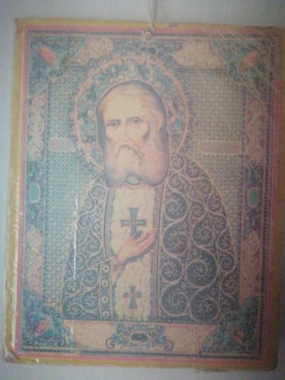 The image displays an Orthodox Christian icon, possibly a printed reproduction, depicting a saint with a white beard and serene expression. The saint is wearing ecclesiastical vestments, including a patterned robe and a vestment with a prominent cross. The background is ornate, featuring intricate geometric patterns and floral motifs in shades of blue, teal, and pink. The icon is framed by a border with decorative elements. The overall impression is that of a religious artifact. There is no discernible information within the image that would indicate a specific location in the city of Yangiyer, Uzbekistan.