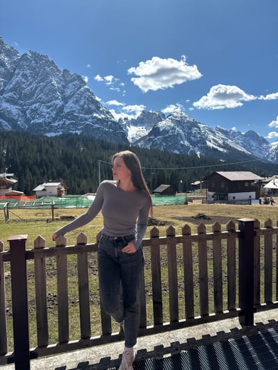A young woman, dressed in a gray long-sleeved shirt and dark jeans, is leaning on a wooden fence in what appears to be Sappada, Italy, given the mountainous backdrop. The snow-capped mountains rise in the background under a blue sky dotted with fluffy white clouds. Houses with brown roofs can be seen nestled among the green trees and grassy fields. The woman is facing towards the mountains, resting her arm on the fence. The scene is bathed in sunlight, suggesting it is daytime. The fence has horizontal wooden slats with dark brown posts.