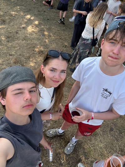 Four people are visible at an outdoor event. Two males and one female take center stage, with a partial view of another person with a beer in the upper right corner. The group stands on dry grass, possibly at a festival or concert. The central female wears sunglasses and a white shirt. One of the males wears a white Stussy shirt and red shorts, holding a red beverage. The other male wears a gray shirt and hat. Wristbands are visible on some of the individuals.