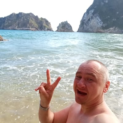 In this selfie, a man stands at the edge of the water on a beach, with dramatic rocky cliffs rising from the sea behind him. He is shirtless and smiling widely, with his mouth open, and holds up his left hand in a peace sign. He is wearing a silver watch on his left wrist. The water is clear and turquoise, with gentle waves lapping at the shore and foam visible where they break. The sky is bright, suggesting it is daytime, likely a clear or partly cloudy day suitable for a beach outing. The jagged, green-tinged cliffs are characteristic of coastal landscapes and suggest a natural, undeveloped area. The scene evokes a feeling of enjoyment and relaxation, possibly on vacation, given the setting and the man's cheerful demeanor. The location is identified as Ribadedeva, Spain, which is known for its rugged coastline and natural beauty. There is no visible text in the image.