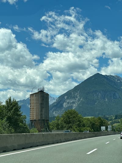 An outdoor, eye-level, medium shot captures a water tower standing amidst a mountainous landscape near the city of Münster, Austria. The sky above is a bright blue, dotted with fluffy white clouds, creating a dynamic contrast with the scene below. In the foreground, a highway stretches out, its grey asphalt surface marked with white lines indicating lanes. A concrete barrier runs along the side of the highway, separating it from the surrounding environment.
To the left of the highway, a lush green forest thrives, its trees forming a dense canopy. Beyond the forest, the imposing structure of the water tower rises tall. The tower itself is cylindrical, with a latticed exterior, and appears to be constructed from wood and metal. It stands on a dark, metallic base, and at its apex, there is a metal railing and a circular antenna-like structure. A ladder is visible on the side of the tower, providing access to its top.
In the background, a majestic mountain range dominates the horizon. The mountains are covered in dense green forests, with rocky outcrops and ridges visible on their slopes. The scale of the mountains dwarfs the water tower, emphasizing the natural beauty of the Austrian landscape. The scene is bathed in natural daylight, with the sun casting shadows and highlighting the textures of the environment. There are no people visible in the image, and the overall impression is one of tranquility and natural grandeur.