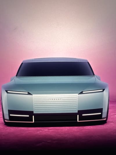 The image displays the front view of a futuristic, light blue concept car with a black grille and thin, illuminated headlights. The car is positioned against a pinkish-purple background. The car's design is sleek and minimalist, featuring sharp lines and a flat front. A subtle "jaguar" logo is visible on the grille. The overall impression is one of a high-tech automotive design study. The image does not provide any information about the location in Sant'Angelo di Piove di Sacco, Italy.