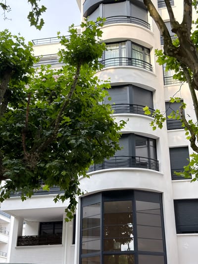 The image shows a modern white building with rounded balconies and dark-paneled windows, partially obscured by the lush green leaves of trees. The building appears to be an apartment complex or office building. The architecture suggests a contemporary design. The surrounding foliage indicates an outdoor setting, likely in a city environment. The bottom part of the image shows a lower section of the building with large glass windows and a white brick facade. Another building, with balconies, is visible in the background on the left. The lighting suggests it is daytime, possibly overcast or late afternoon, as there are no harsh shadows. The overall impression is one of urban living with elements of nature integrated into the scene. The building in the foreground is located in Maisons-Alfort, France.