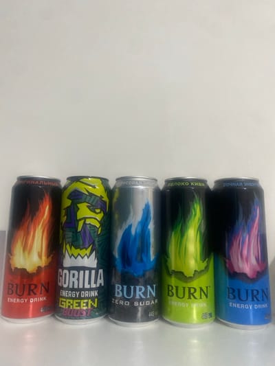 Five energy drink cans are stacked horizontally. From top to bottom: a blue can with pink flames labeled "Burn Energy Drink," a green can with green flames labeled "Burn Energy Drink," a silver can with blue flames labeled "Burn Zero Sugar," a green and yellow can labeled "Gorilla Energy Drink Green Boost," and a red can with orange flames labeled "Burn Energy Drink." Russian text labels each of the top three cans.