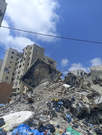 The image depicts the aftermath of destruction in the city of Jabalia, Palestinian Territories. A multi-story building, heavily damaged and partially collapsed, dominates the scene. Debris from the building, including concrete slabs, rebar, and rubble, is piled high in the foreground. Scattered amongst the rubble are various forms of trash, including plastic bags, bottles, and cans, suggesting a significant amount of refuse has accumulated in the area.
In the background, more damaged structures are visible, with broken windows and exposed walls indicating widespread destruction. The sky above is a clear blue with scattered white clouds. Power lines are strung across the frame, some appearing to be intact while others are severed or hanging precariously. The overall impression is one of devastation and neglect. There are no people or animals visible in the image.